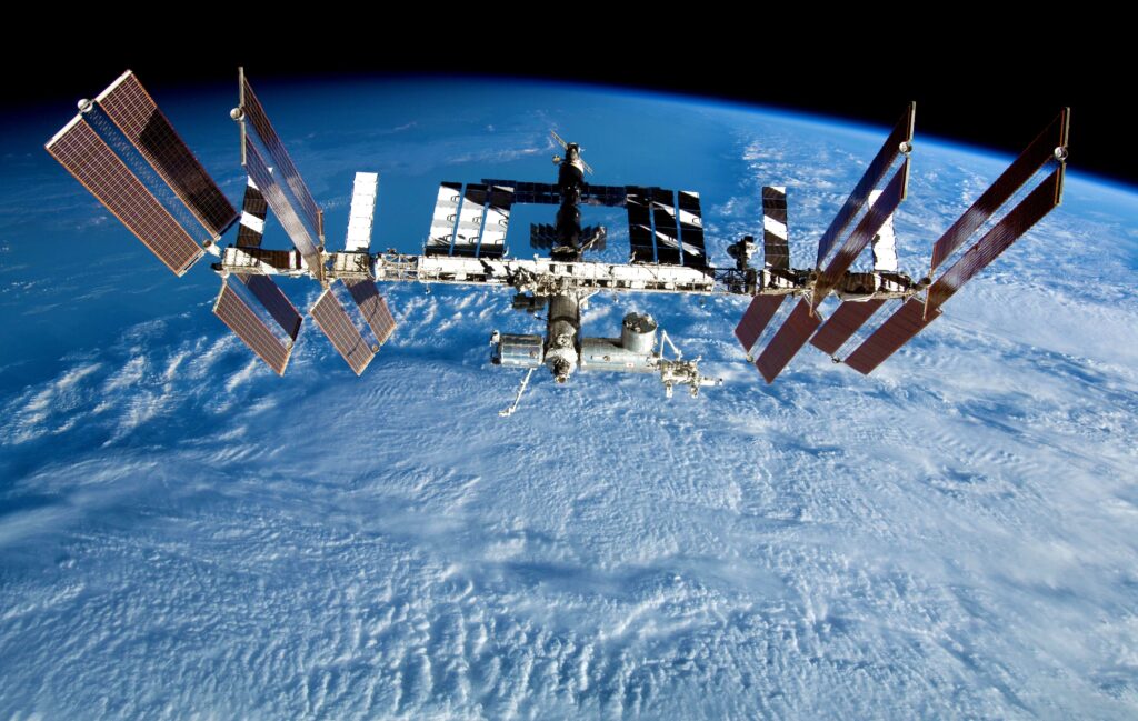 A silver and gray space station floats above the blue and white cloud-covered Earth.