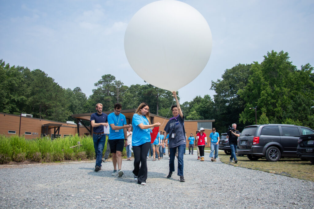 Two people are carrying a large white balloon aloft while walking down a gravel path.