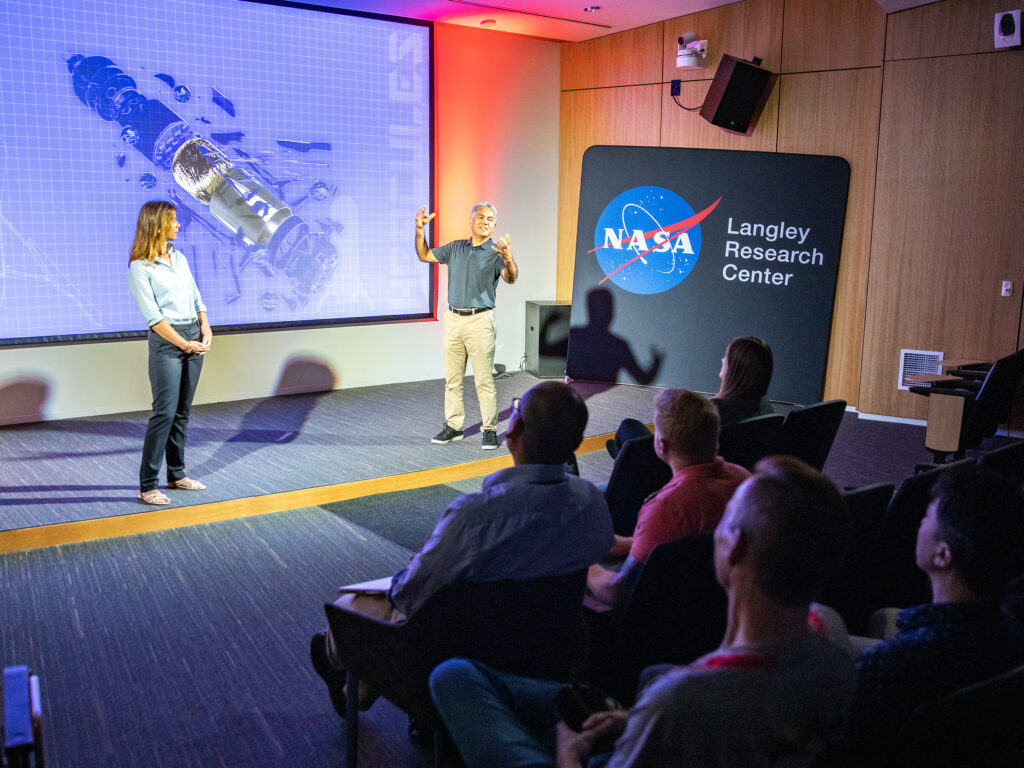 Two aerospace engineers are standing on a stage, talking animatedly in front of an audience in a dark theatre. On the screen behind the speakers is an exploded view of a rocket, showing the parts of the system. To the right of the stage is a display that shows the NASA logo and the words "Langley Research Center"