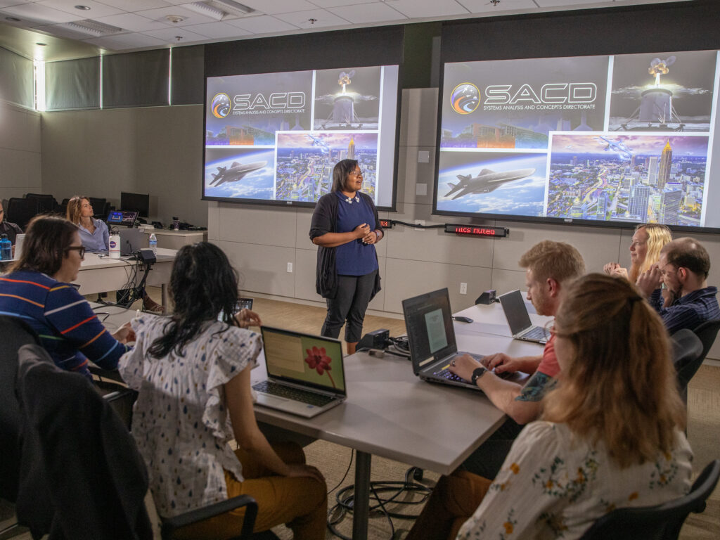 A group of aerospace engineers are sitting at long, rectangular tables with their laptops open while one engineer stands at the front of the room in front of two screens that show the directorates logo and artistic renderings of air and space vehicles.