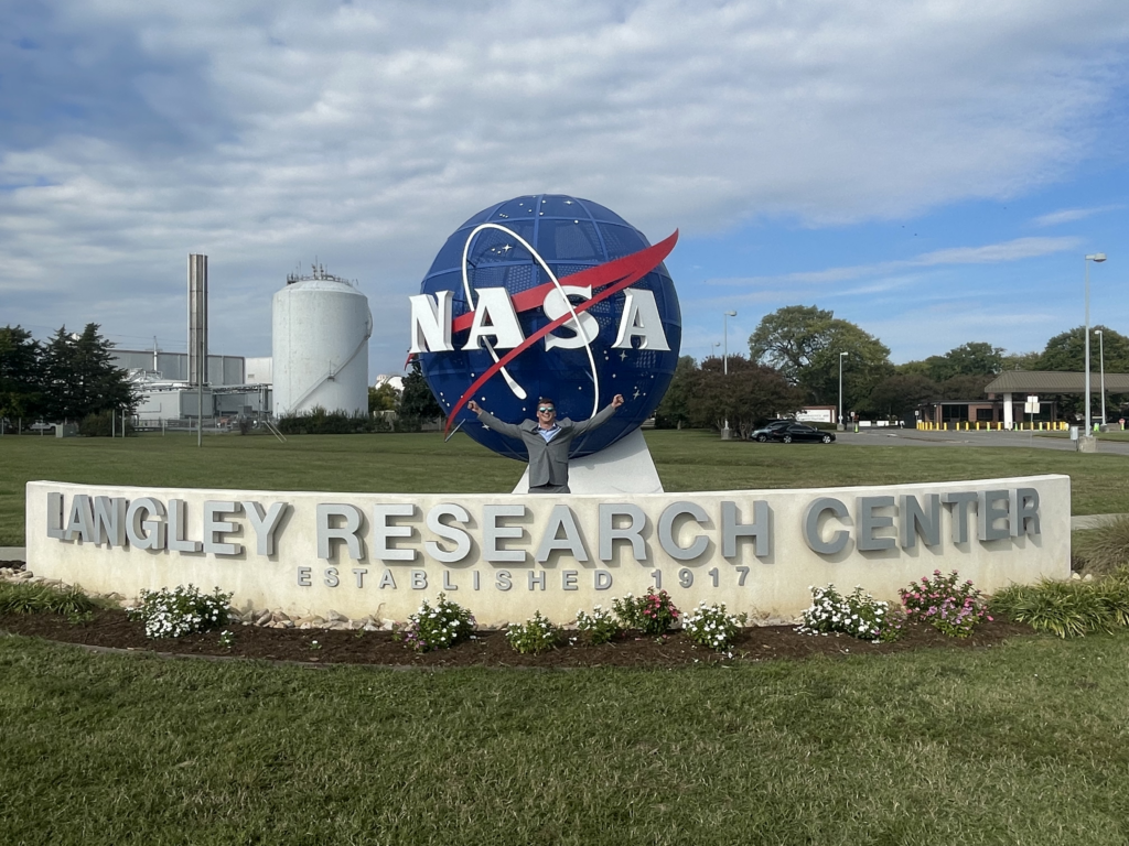 A smiling man with outstretched arms stands at the entrance gate for Langley Research Center. A statue of the NASA logo is positioned behind a large brick wall that reads NASA Langley Research Center.